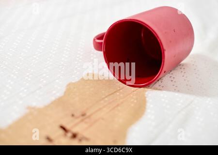 Red mug tipped over, spilling coffee on white fabric. Coffee granules scattered across surface. Bright lighting highlights contrast between red mug an Stock Photo