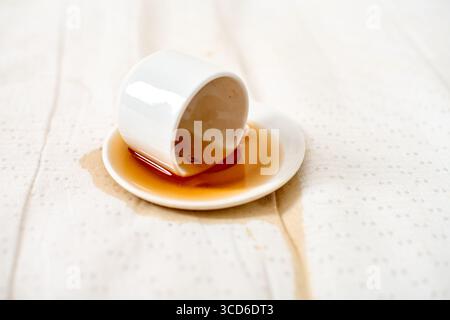 White cup overturned, coffee spilled on saucer and fabric. Soft natural lighting highlights liquid trail, creating casual yet dramatic contrast. Simpl Stock Photo