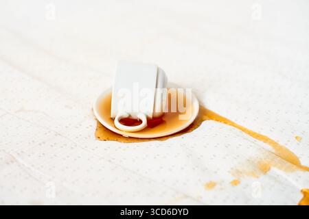 Upside-down white mug rests on light tablecloth, coffee spilling out. Brown stain spreads across textured fabric, creating contrast. Overhead perspect Stock Photo