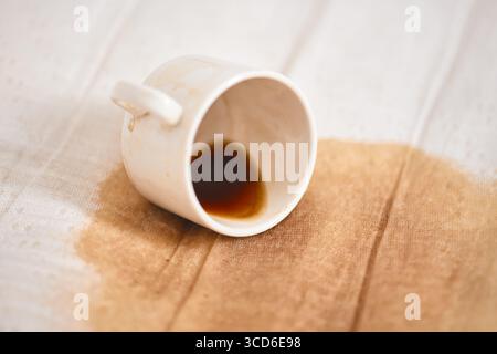 White ceramic cup resting on side with small coffee puddle on light fabric. Simple composition emphasizes spill, creating contrast between dark coffee Stock Photo
