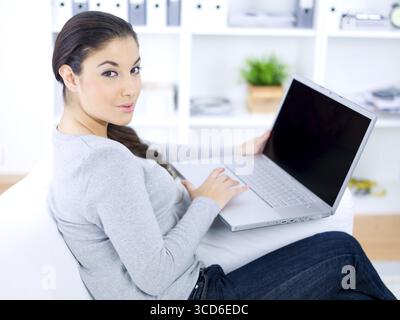 Woman using a laptop in the living room as she sits on a comfortable sofa turning to look at the camera with a smile Stock Photo