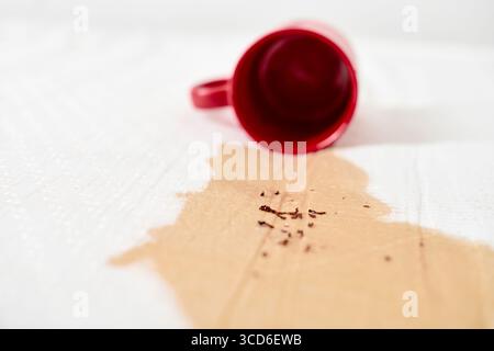 Red mug tipped over, spilling coffee on white fabric. Coffee granules scattered across surface. Bright lighting highlights contrast between red mug an Stock Photo