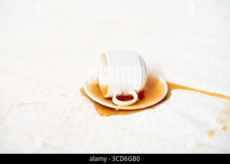 Upside-down white mug rests on light tablecloth, coffee spilling out. Brown stain spreads across textured fabric, creating contrast. Overhead perspect Stock Photo