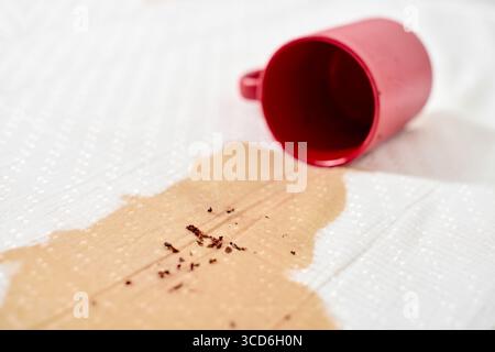 Red mug tipped over, spilling coffee on white fabric. Coffee granules scattered across surface. Bright lighting highlights contrast between red mug an Stock Photo