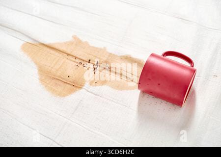 Red mug lies on side, spilling coffee onto white fabric. Light coffee stain spreads, creating splash effect. Contrasting colors highlight spills promi Stock Photo