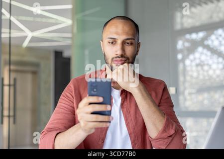 A focused individual holding a smartphone while seated in a contemporary office setting. Stock Photo