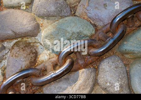 Rusty chain links stretch across a rugged stone path, blending smooth and rough stones under natural light, highlighting the contrast between organic Stock Photo