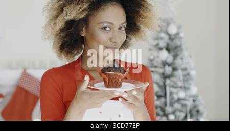 Young African cook with a freshly baked Christmas cake in her hand closing her eyes in bliss as she enjoys the aroma Christmas tree background Stock Photo