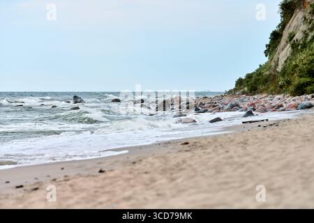 Dynamic waves crash against rocky shoreline under soft daylight. Sandy beach foreground contrasts with textured cliffside. Natural baltic beauty in se Stock Photo