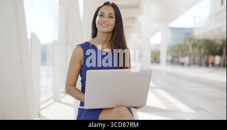 Thoughtful young businesswoman working outdoors enjoying the fresh air sitting on a bench on an urban promenade using her laptop computer Stock Photo