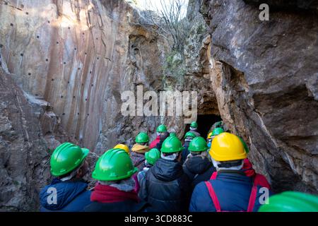 A group of people wearing green and yellow helmets entering a cave or tunnel, surrounded by rocky walls Stock Photo