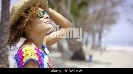 Pretty adult female in reflective yellow sunglasses and straw hat outside on beach looking upward with copy space Stock Photo
