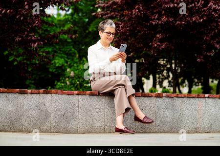 Mature professional woman using a mobile phone while sitting outdoors on a sunny day in a business casual outfit Stock Photo