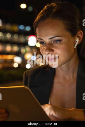 Portrait of modern female entrepreneur with wireless earbuds using digital tablet in night city Stock Photo