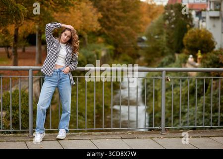 Young woman in trendy autumn outfit posing on a European bridge with golden foliage and a historic cityscape in the background. A stylish young woman Stock Photo