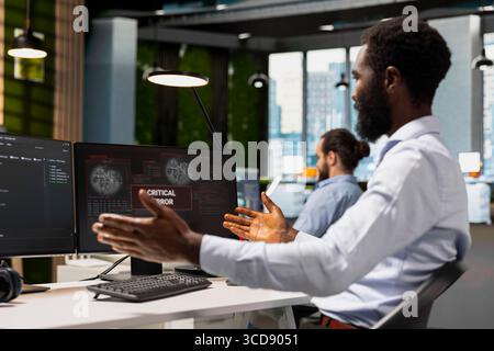 Software developer in office surprised by critical error warning popup, struggling to fix it. African american man troubleshooting error notification on PC screen interrupting workflow Stock Photo