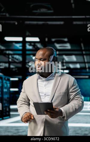 Admin listening music in data center, using tablet to oversee infrastructure mainframes. Worker wearing audio headphones walking in server farm, using tablet to supervise hardware rigs Stock Photo