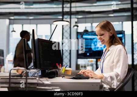 Cheerful junior dev writing code, developing machine learning APIs. Happy young female engineer working in office, troubleshooting software errors using artificial intelligence tech Stock Photo