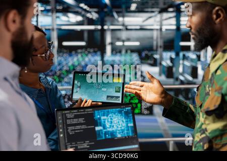 Military technicians in secure data center monitoring AI tech systems. Black army operator in server hub ensuring stable infrastructure for machine learning defense operations. Stock Photo