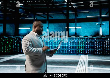 Developer in data center updating systems using laptop, protecting mainframes from damaging software errors. Server hub IT expert using notebook, looking for malfunctions causing gear to slow down Stock Photo