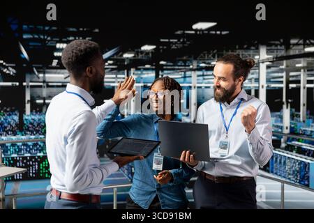 Multiethnic confident team sharing a high five to celebrate achievement on server farm industrial platform, cheering to their professional collaboration. Pleased successful engineering team. Stock Photo