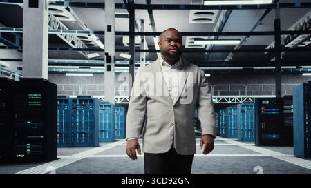 Smiling engineer walking in data center, checking hardware for system stability. Happy employee overseeing server farm, configuring infrastructure to optimize performance, camera A Stock Photo
