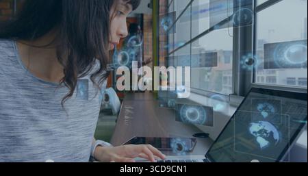 Typing female professional working on laptop at office under large windows, with holographic data Stock Photo