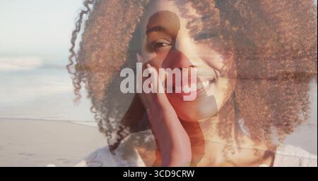 Smiling woman in white blouse touching cheek on beach, with man in coat walking silhouette Stock Photo