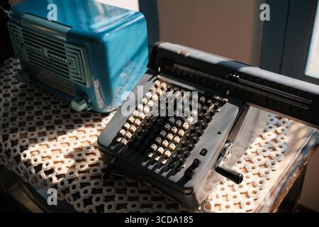 Vintage mechanical calculator and retro blue radio on a lace-covered wooden table in warm natural sunlight. Stock Photo