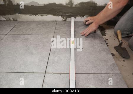 A building contractor is installing large square slip resistent tiles on a bathroom floor using a spirit level and trowel to apply mortar. Stock Photo
