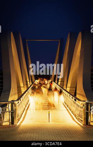 Night view of the Wave of Azov pedestrian bridge in Kyiv, Ukraine, with blurred silhouettes of people in motion under warm lights against a deep blue Stock Photo