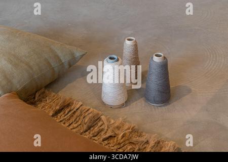 Three spools of thread in neutral colors placed on a textured surface with fabric cushions nearby. The scene conveys a cozy, creative atmosphere. Stock Photo