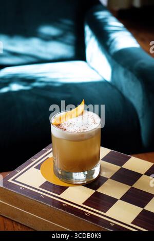 Whiskey sour cocktail served in a short glass with a creamy foam top,  Drink placed on a wooden chessboard with a cozy armchair Stock Photo