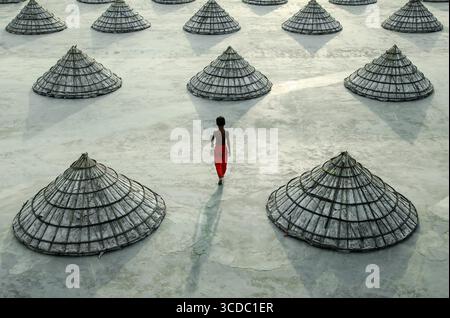 Brahmanbaria, Bangladesh - 08 June 2021: View of a solitary figure in red walking amid a sea of neatly arranged, conical structures under the vast sky. Stock Photo