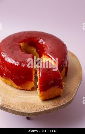 A fresh homemade bundt cake topped with a vibrant red berry glaze, served on a rustic wooden cake stand against a pink background with a slice cut out Stock Photo