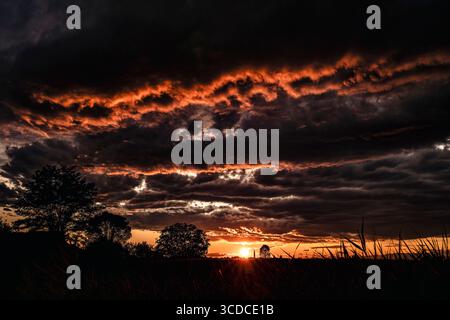 Fiery sunset painting the sky with vibrant orange hues behind dark clouds over a field, creating a dramatic and moody landscape Stock Photo