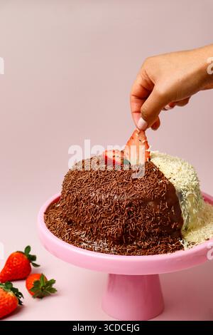 A hand places a fresh strawberry slice on a Brigadeiro cake, a traditional Brazilian dessert, which is half-covered in chocolate sprinkles Stock Photo