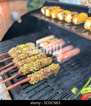 Lamb kebabs, sausages and corn on cob, cooking on an outdoor grill in summer. In the foreground the Adana lamb kebabs on cinnamon sticks. Stock Photo