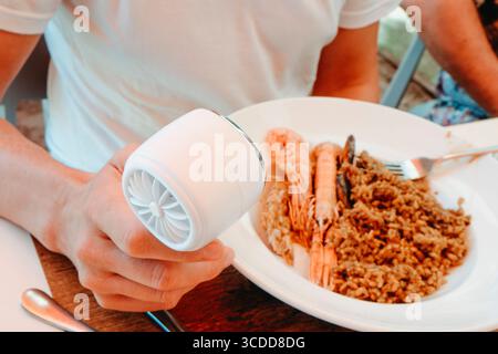a man in a white t-shirt cools himself with a small handheld fan while sitting at an outdoor restaurant table in front of a plate of seafood rice on a Stock Photo