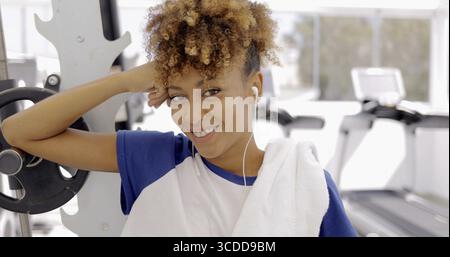 Portrait of young curly model in sportive clothing posing in headphones and with towel on neck smiling happily at camera on background of gym Stock Photo