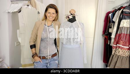 Young smiling female tailor posing with mannequin in dress and smiling at camera in parlour Stock Photo