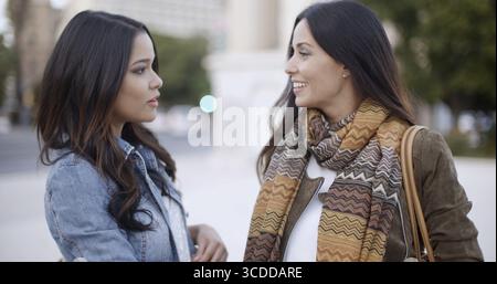 Two stylish smiling young women standing facing each other chatting outdoors in a town square head and shoulders in winter fashion Stock Photo