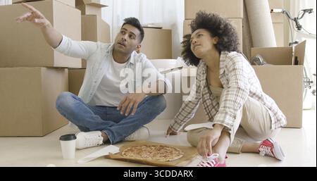Young couple eating pizza on the floor in front of a stack of brown packing boxes as they move house and chatting pointing to something off screen Stock Photo