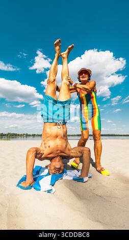 Elderly man doing assisted headstand on beach towel with smiling woman helping Stock Photo