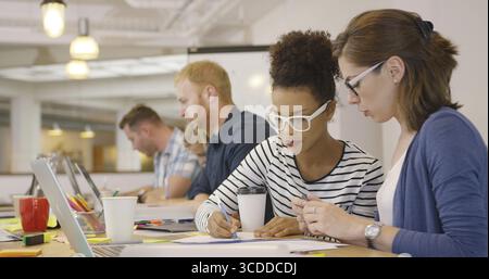 Two young female colleagues working on one document while sitting at long table with male coworkers Stock Photo