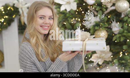 Smiling blond woman posing with Christmas wrapped gift box next to pine tree Stock Photo