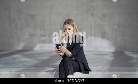 Positive blond female manager in smart casual clothes sitting on concrete bench and browsing social media on smartphone Stock Photo