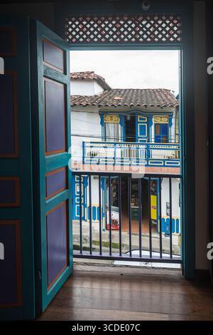 View through opened door to the opposite side of the street: Colorful house with balcony and shop. Salento, Quindío Department, Colombia Stock Photo