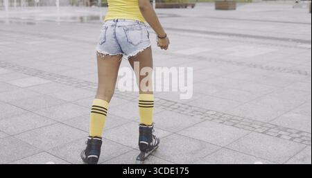 Crop back view of woman in denim shorts and knee stockings skating in rollers on background of street pavement Stock Photo
