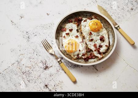 Two fresh fried eggs with crunchy crisp bacon served on rustic plate. Fork and knife on sides. Top view Stock Photo